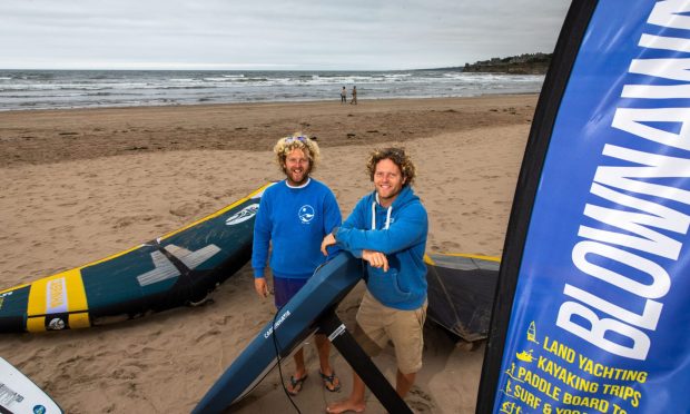 Image shows: Guy and Jamie McKenzie of Blown Away on West Sands beach, St Andrews. Both men, who are twins, have blond curly hair and are wearing blue hoodies and are setting up their kites and foilboards for some wing foiling in the sea.