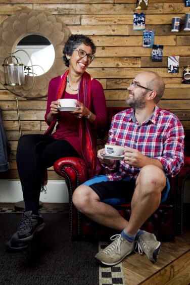 Image shows: Menopause cafe founders Rachel Weiss and Andy Sanwell. The couple are sitting on a red leather sofa and drinking coffee at a Menopause Cafe in Perth's Blend coffee shop.