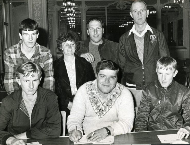 Jocky Wilson sits at a table and signs autographs for fans, who gather round him to pose for a picture