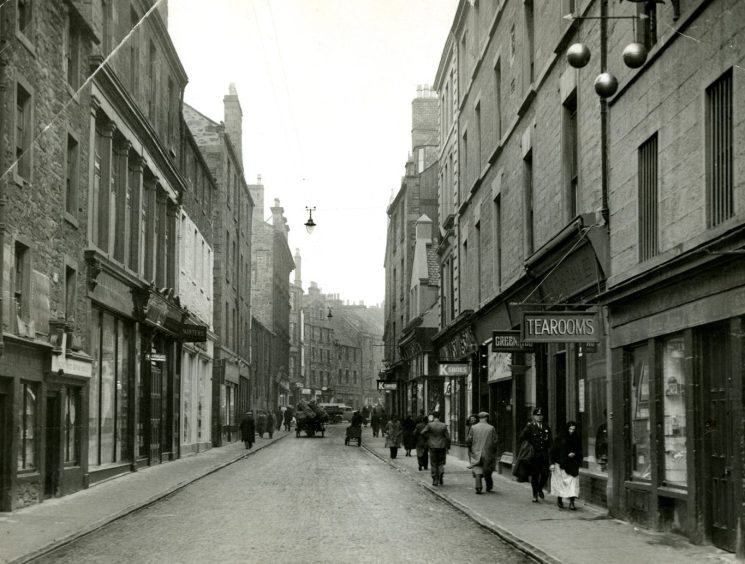 People walk on the pavement as a horse-drawn cart makes its way along a street in the Overgate.