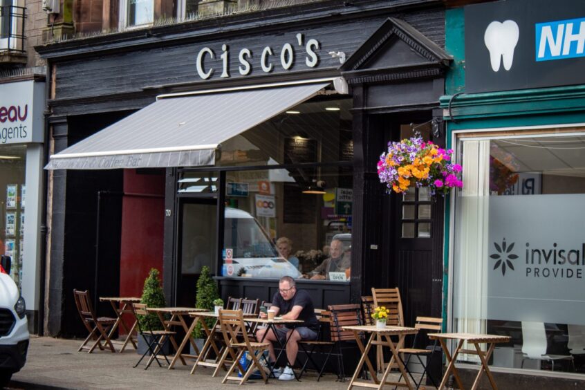 A man sits at a table outside Cisco's cafe on Port Street in Stirling, while people dine inside, visible through the window.