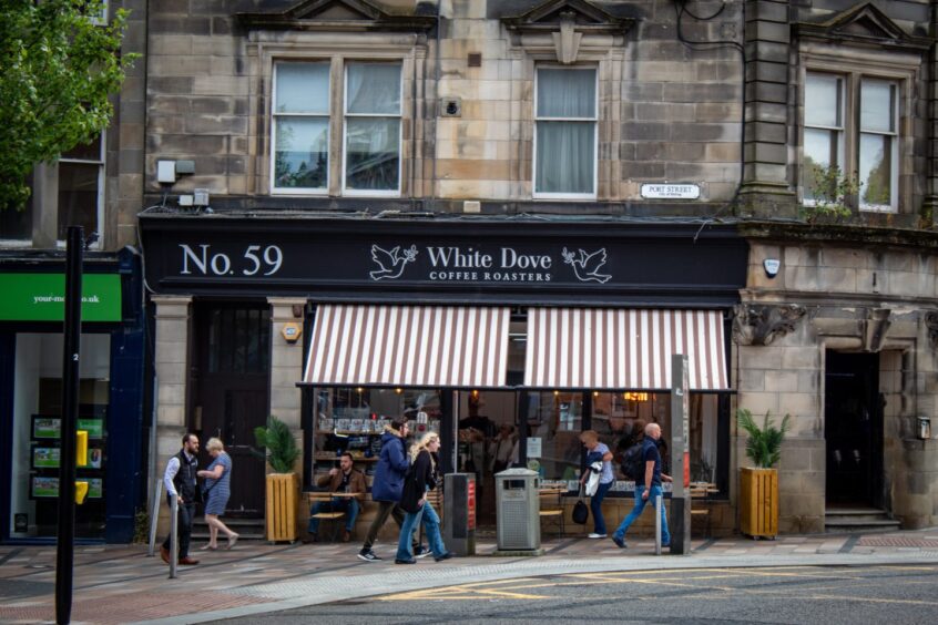 People walk past the exterior of White Dove Coffee Roasters cafe on Port Street in Stirling, with striped awnings and tables outside.