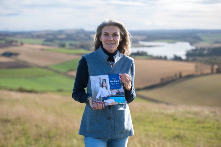 Anona Gow holding her new book on the hill above Pitscandly Farm, Forfar. There are beautiful views behind her of farmland and a loch.