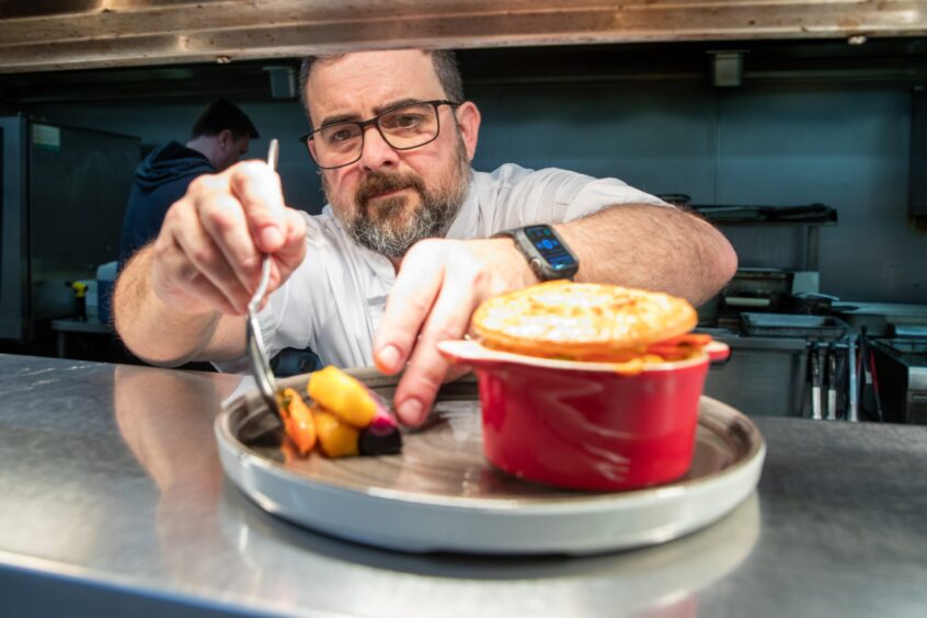 The Meadowpark's head chef Jesse Spears dishing up heritage carrots onto a plate containing a red ramekin of lobster pie. Jesse has a dark short beard and dark-rimmed glasses.