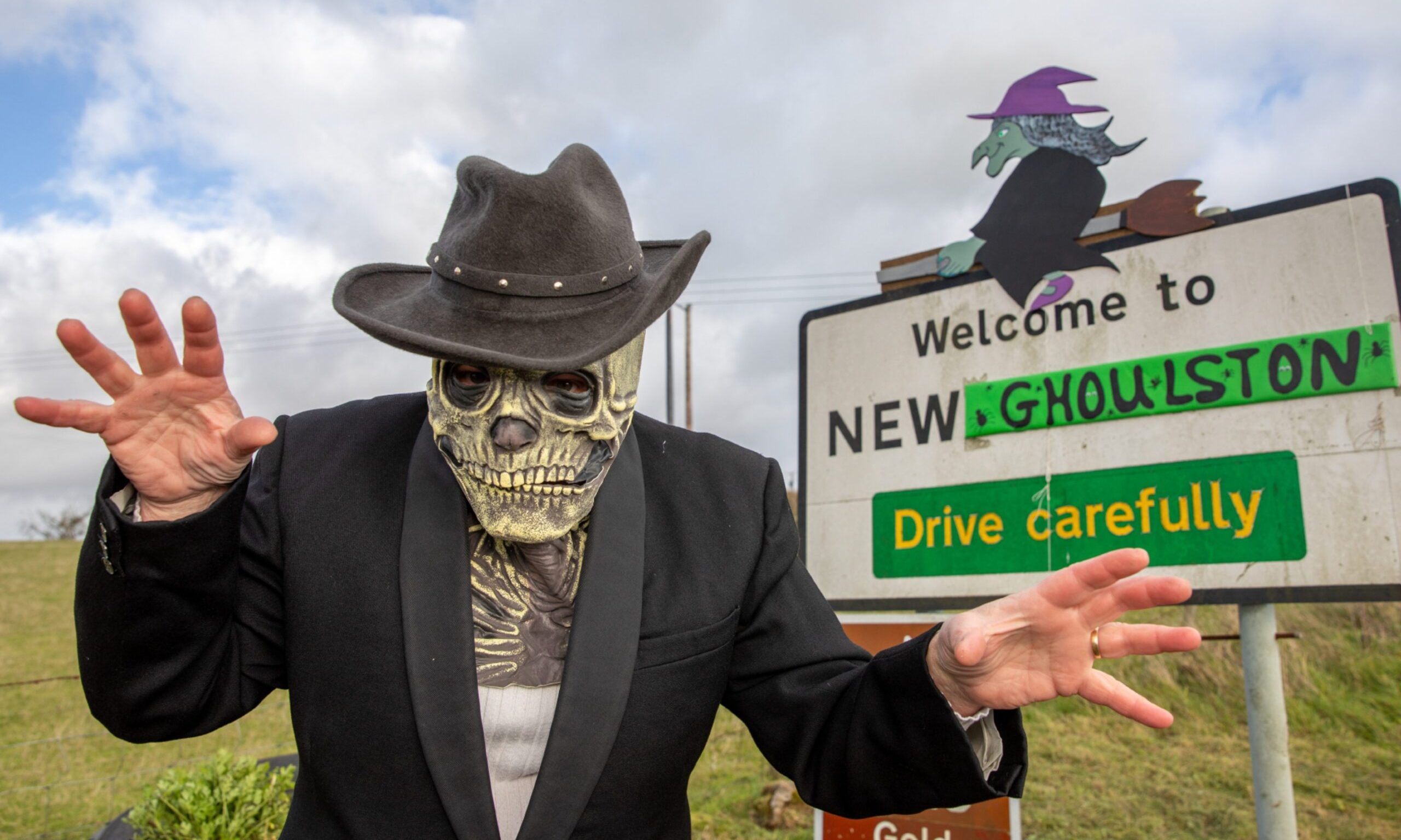 Image shows the Welcome to New Gilston signpost outside the Fife village. The village has been renamed New Ghoulston for Halloween and the signpost has been changed which Ghoulston written over Gilston and a witch riding on top of the sign. A scary character wearing a skeleton mask and black suit and hat and standing in a scary pose.