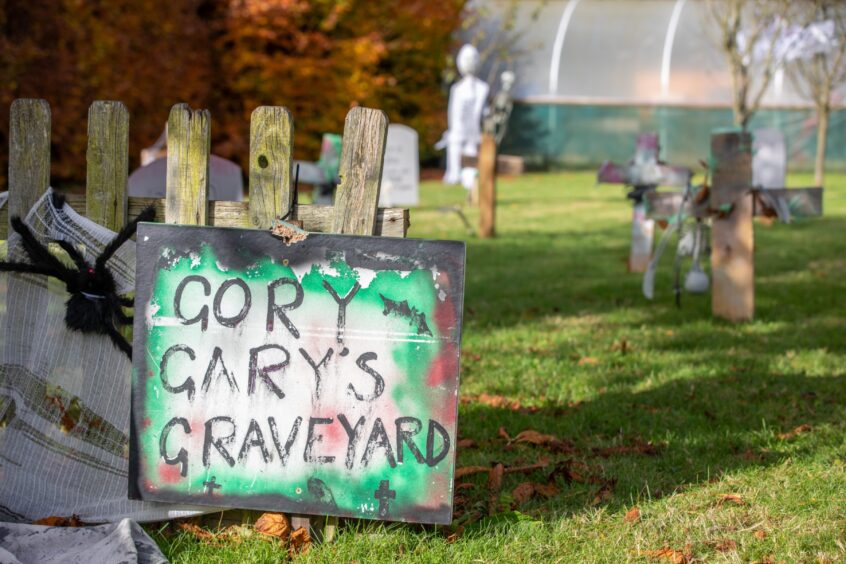 Image shows: a handmade scary-looking sign reading Gory Gary's Graveyard. The sign sits at the entrance to a Halloween graveyard created at New Gilston Village Hall.