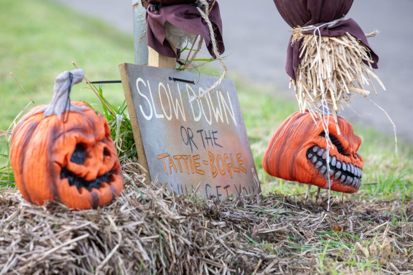 Image shows: a spooky sign in New Gilston. The sign reads Slow Down or the Tattie Bogle will get you! There is a scarecrow above the sign and scary pumpkin-heads on either side of it.