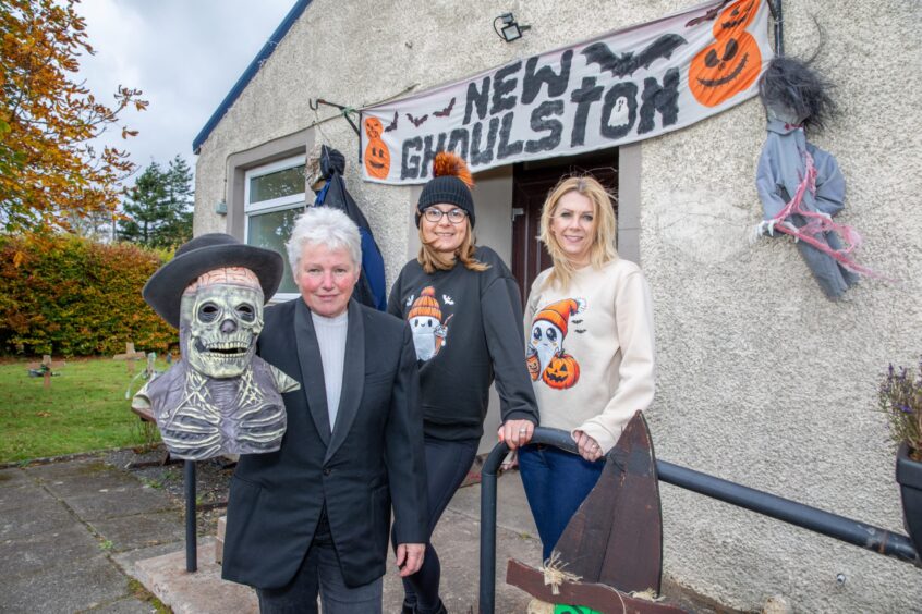 Image shows: three women outside a village hall in Fife. Dianne Penrice, Kim Coupar and Leanne Law are wearing halloween jumpers and outfits and standing under a banner that reads New Ghoulston.