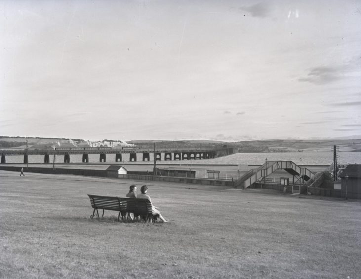 two people sit on a bench with a view of Magdalen Green Station as a train goes over the Tay Bridge 