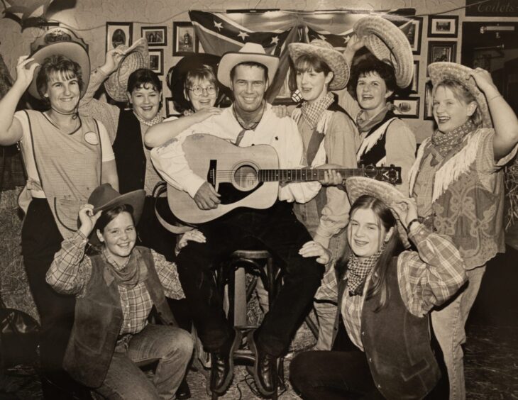 Black and white photo of man dressed as cowboy with guitar surrounded by women raising cowboy hats