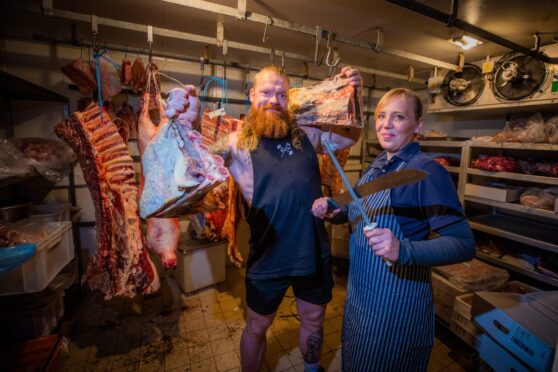 John Beattie holding side of beef next to Wendy Donald sharpening knives in her butcher shop cold room