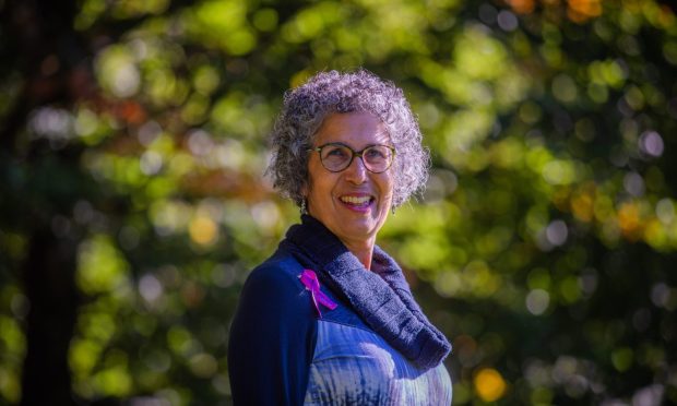 Image shows: Menopause awareness campaigner Rachel Weiss who is wearing a purple Menopause Cafe support ribbon. Rachel is standing smiling at the camera and has grey curly hair and wears glasses. She is pictured on a sunny September day with trees in the background.