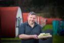 Pierre Leger holding tray of Strathearn Cheeses in front of row of POW camp huts