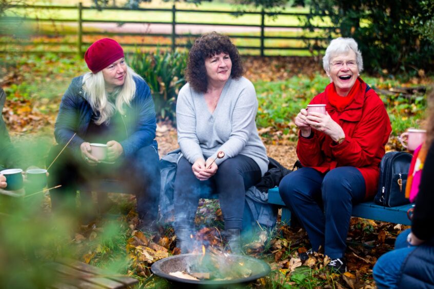 Image shows: a group of walkers enjoying hot drinks around a fire pit after an autumn walk. Rosie Henshaw, Jacky Moug and Jean Kemp are smiling and enjoying the post-walk chat.