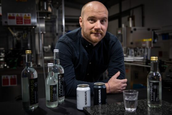 Vivimus Water founder Darren Peattie leaning on a counter in his factory with cans and bottles of his water.