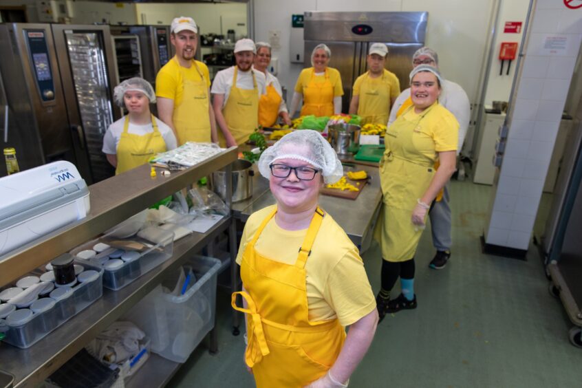 Chloe Hutchison in the working kitchen of The Sunshine Kitchen smiling with her team, all wearing bright yellow aprons.