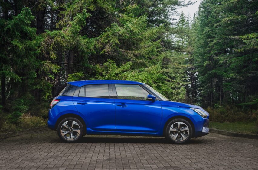 A blue Suzuki Swift seen in profile with woods in the background