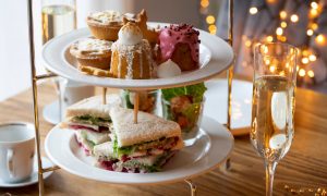 A magnificent looking festive afternoon tea with mince pies, tasty looking mini cakes and sandwiches. next to a flute of champagne.