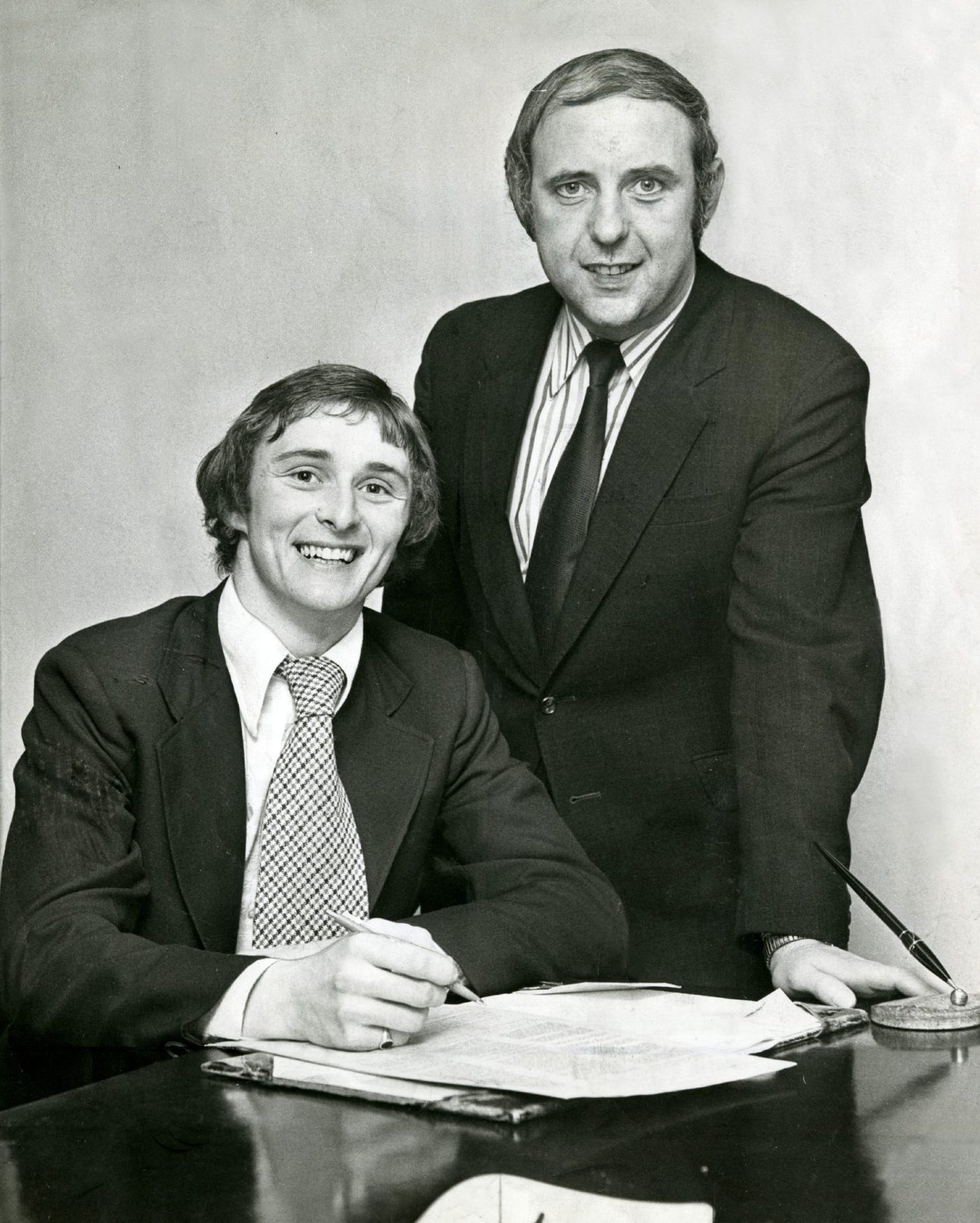 Both wearing suits, Jim McLean watches Hegarty sign on the dotted line on November 12 1974.