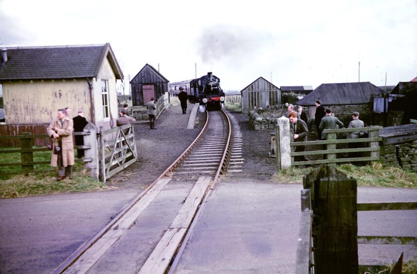 people look on at a railway crossing as the double-headed rail tour reaches Carmyllie in April 1962