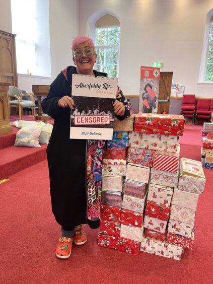 Zoe Dark smiling as she holds a calendar in front of a pile of shoeboxes inside a church hall