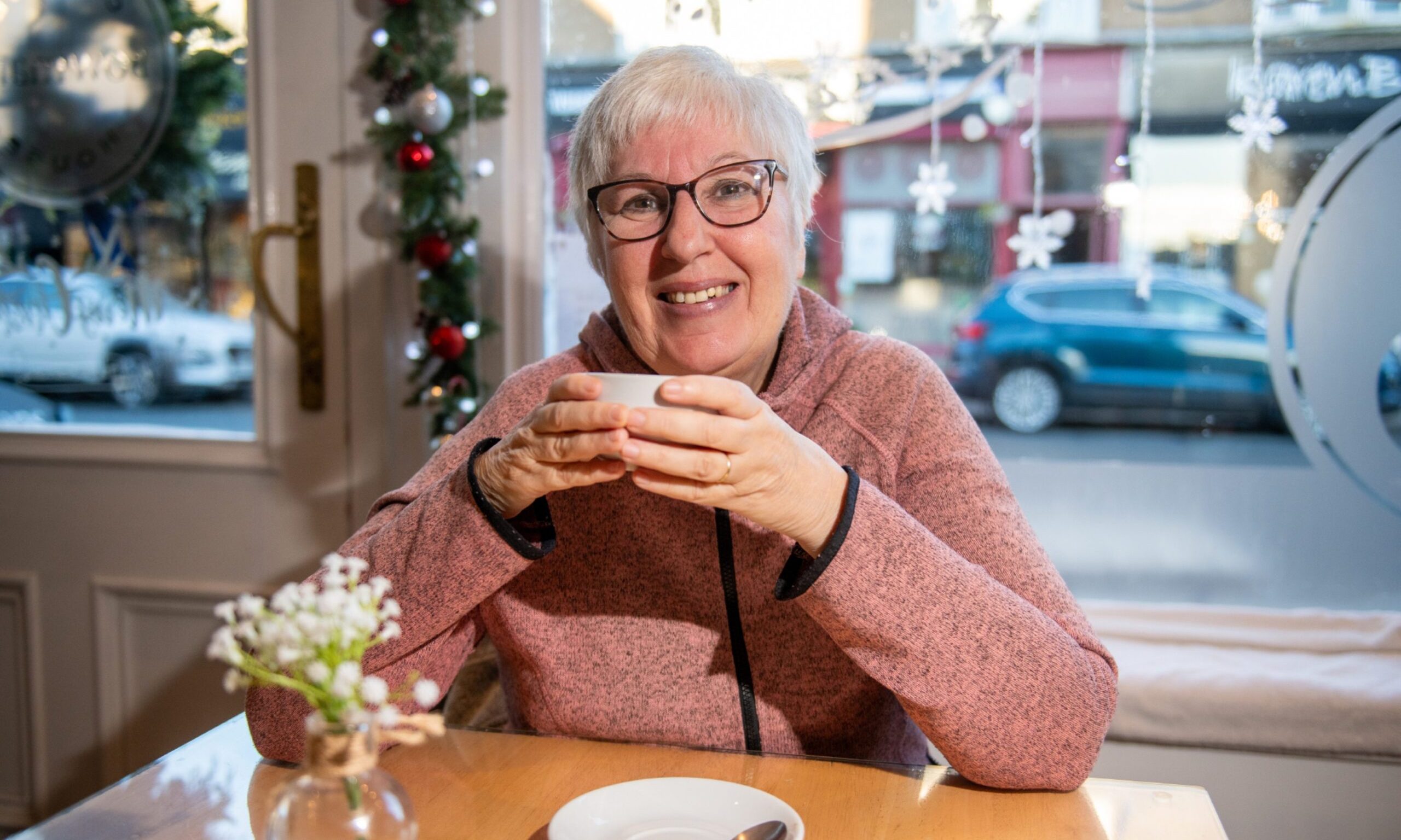 Image shows: Travel writer Helen Moat enjoying a cup of coffee in a cafe in Broughty Ferry. Helen is sitting at a table in the window of Bowmans smiling and holding a cup in her hand. She has short white hair and glasses and is wearing a pinky coloured fleece.