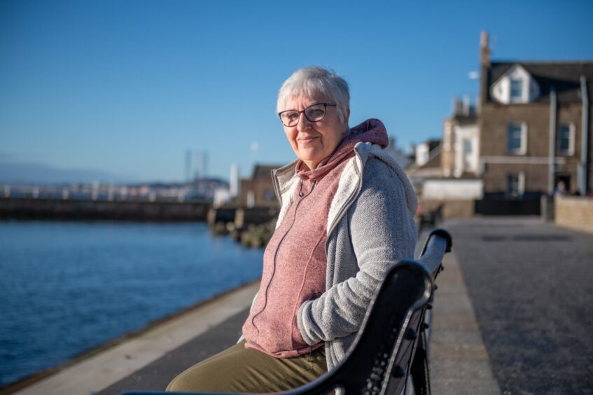 Image shows: Helen Moat, author of While the Earth Holds Its Breath, sitting on a bench on the seafront at Brought Ferry. It is a cold, crisp winter's day and the sky and sea are blue.