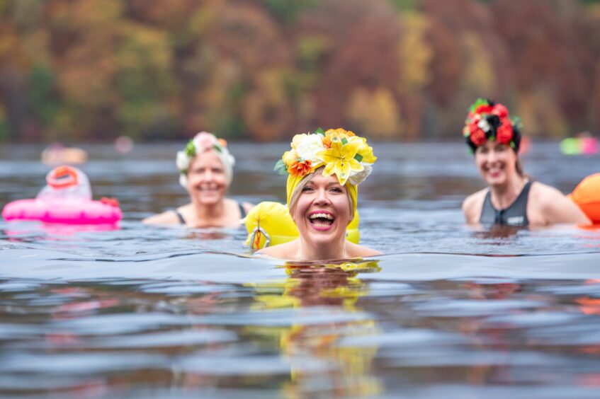 Image shows: three women swimming in Loch Faskally in Perthshire. The women are wearing bright swimming hats and wide smiles. The water is still and the backdrop shows autumn leaves on the trees.