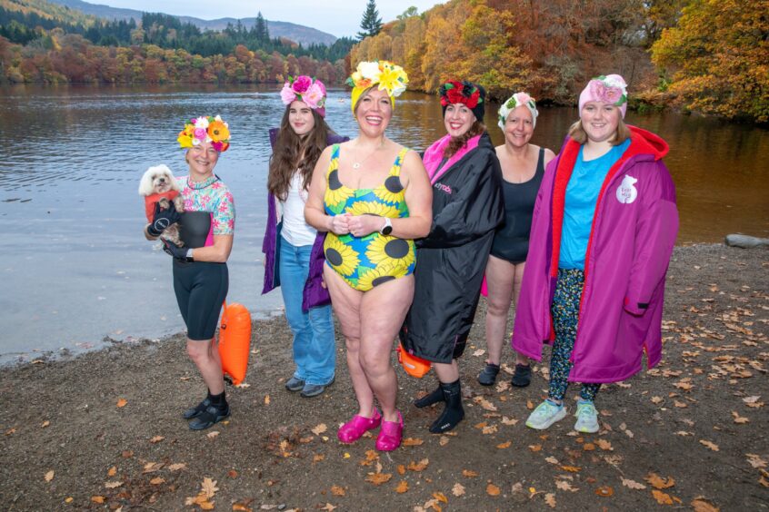 Image shows: a group of women on the shore of Loch Faskally, Pitlochry. All of the women are wearing flamboyant swimming hats decorated with colourful flowers. In the centre of the image, Stephanie Scott is wearing a swimming costume with bold sunflower pattern and a swim hat with yellow flowers.