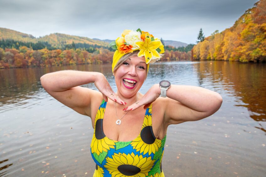 Image shows: Stephanie Scott wearing a bright yellow swimming cap decorated with flowers and a swimsuit with sunflowers on it. She is smiling and posing for the camera. Loch Faskally is in the background in full autumn colour.