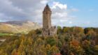 The stone gothic National Wallace Monument tower sits on a hill, flanked by trees with autumn foliage, and a blue sky in the background