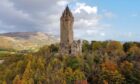 The stone gothic National Wallace Monument tower sits on a hill, flanked by trees with autumn foliage, and a blue sky in the background