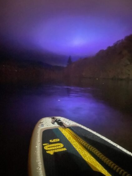 The tip of a paddleboard on Loch Fascally with a purple-tinged sky lit up by the Enchanted Forest illuminations, surrounded by trees.