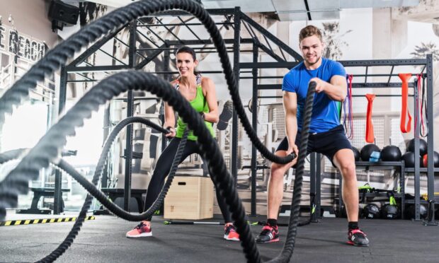 A woman in a green top and a man in a blue top work out in a gym using black battle ropes. In the background, other gym equipment including medicine balls is visible