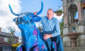 David McCann kneeling next to brightly painted cow sculpture beside Murray Fountain in Crieff