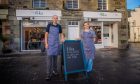 Elizabeth and Jamie Grant stand outside their revamped cafe Fika in Aberfeldy. They wear blue aprons and stand next to a blackboard sign for their new cafe.