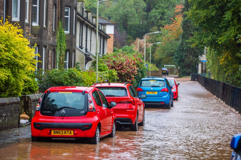 Cars parked outside houses with water pouring down street and sandbags at garden gates