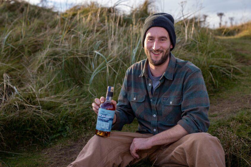 David Taylor of East Neuk Spirit Co. sits in the grassy dunes at Elie with a bottle of Dorchadas Washed Ashore single cask rum.