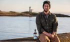 East Neuk Spirit Co. co-owner David Taylor sits on the wall with the lighthouse at Elie behind him and a bottle of his single cask rum next to him.