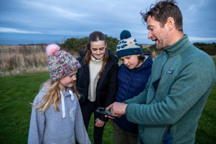 Image shows: Rory Fyfe of Rewilding Kinkell showing the virtual fence system that he controls from his mobile phone to three children who have come to meet the Highland Cows.