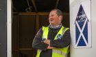 Image shows: Men's Shed founder Henry Paul. Henry is leaning against the door of the Men's Shed building. He is wearing a branded high-vis vest and smiling. The door has a Men's Shed logo which is an illustration of a building in the saltire colours.