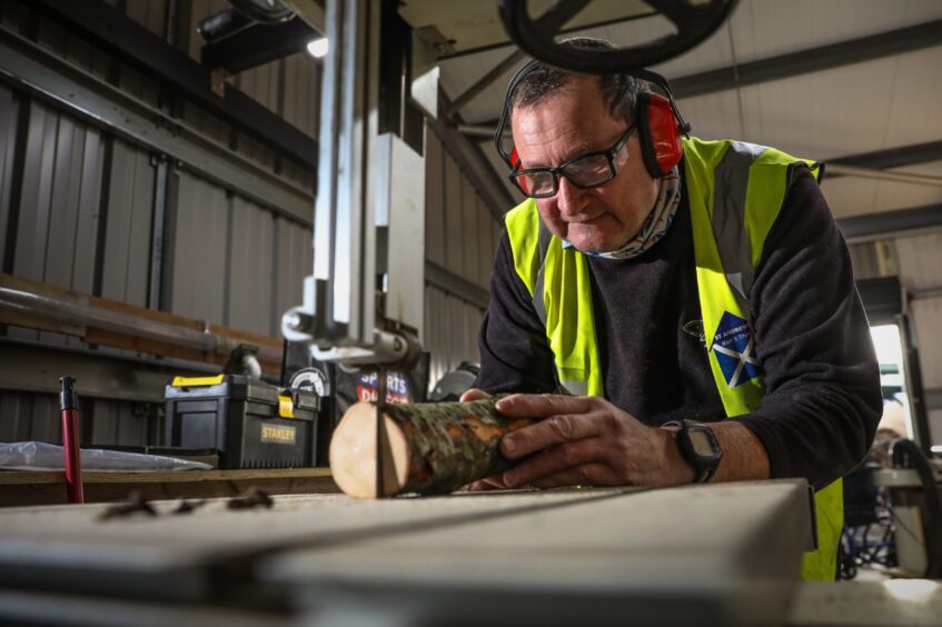 Image shows a man cutting wood in a workshop. He is wearing safety glasses, high vis vest and ear defenders.