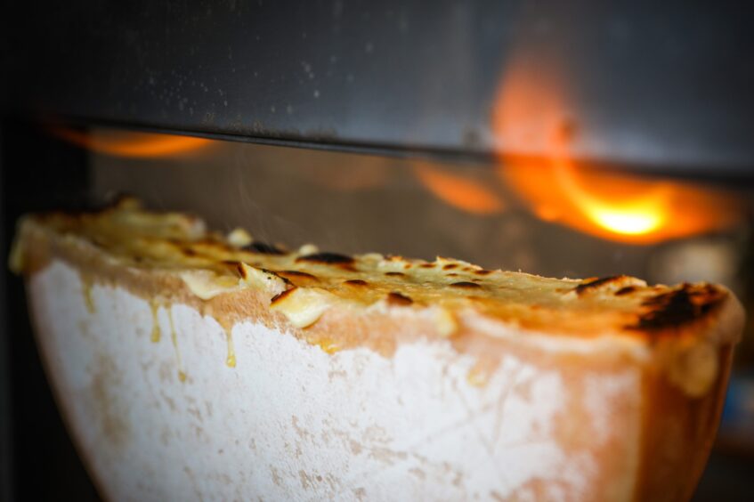 A half wheel of raclette melting under a gas-powered raclette machine flame.