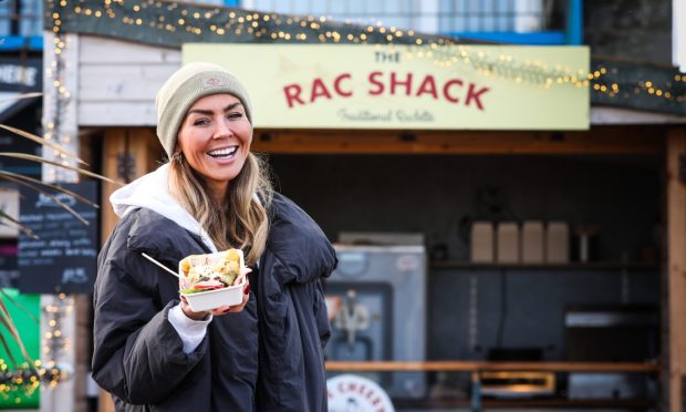 Kate Carter-Larg outside the new food outlet The Rac Shack, holding a portion of raclette. She is smiling and wearing a beanie hat and a black puffer jacker.