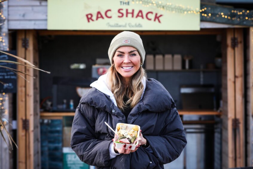 Kate smiling and facing the camera outside The Rac Shack, holding a portion of raclette.