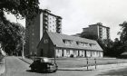 a car turns a corner in front of a row of houses, with the Dryburgh multis rising in the background