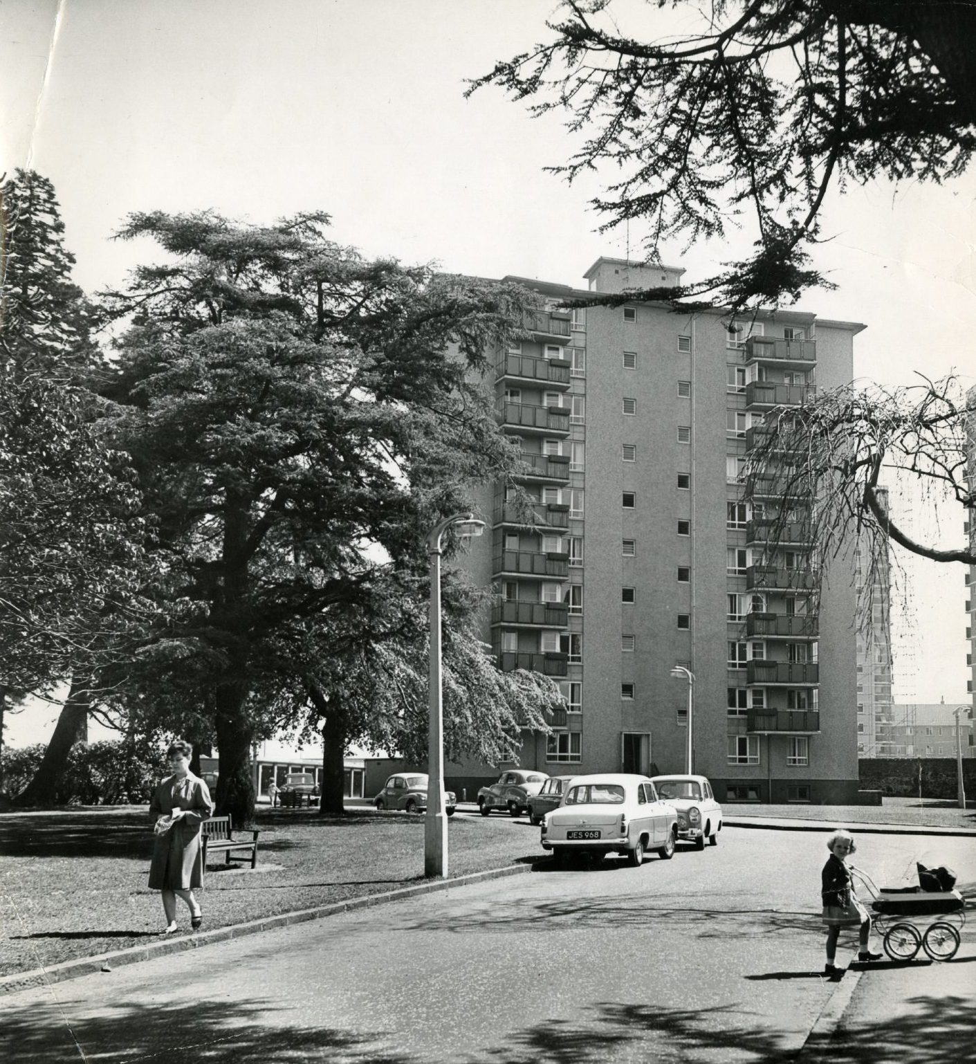 a young girl pushes a pram and woman walks on grass outside the multi storey flats at Dryburgh Gardens in 1964.