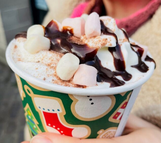 A close-up of the top of a hot chocolate with marshmallows, cream and chocolate sauce in a festive cardboard cup.