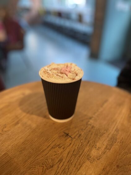 A paper cup of hot chocolate sitting on a table with a cafe and window in the background.