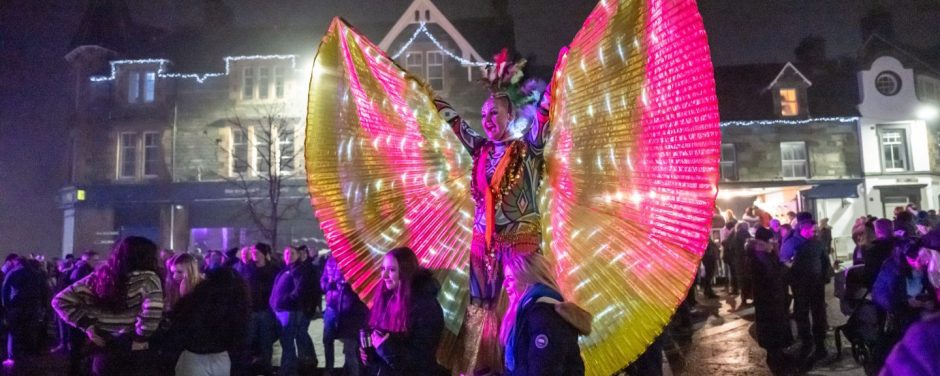 A carnival dancer is lit up and displaying coloured wings to a crowd.